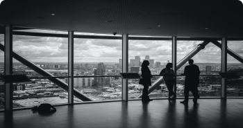 Three people standing and talking in front of large windows overlooking a city skyline with cloudy skies. The environment is a modern office space with an open and professional atmosphere. No visible text in the image. The emotional tone is collaborative and focused.
